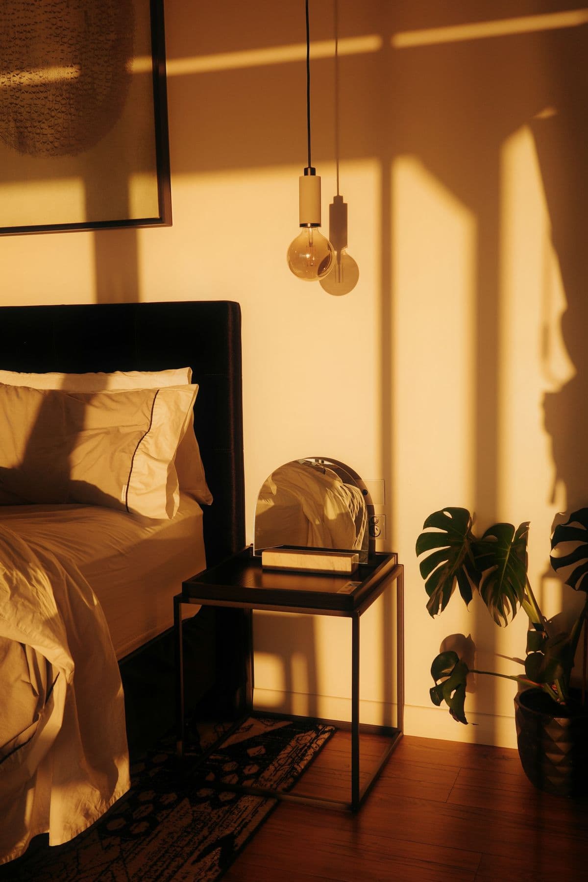 A quiet bedroom in warm evening light, with a made bed, a bedside table and a plant.